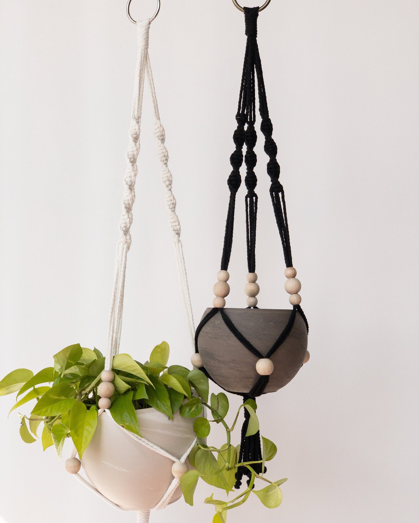 Two macrame plant hangers. One is white, one is black. Each have 3 spiral straps with adjustable beads to hold the pots. The pot on the left has a neon pothos.
