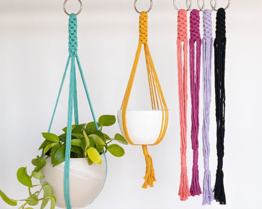 macrame plant hangers in various colors: aqua, yellow, coral. hot pink, lavender and black. They have an intricate tight knot pattern at the top that opens up to straight straps to hold the pot. They have a tassel at the bottom.