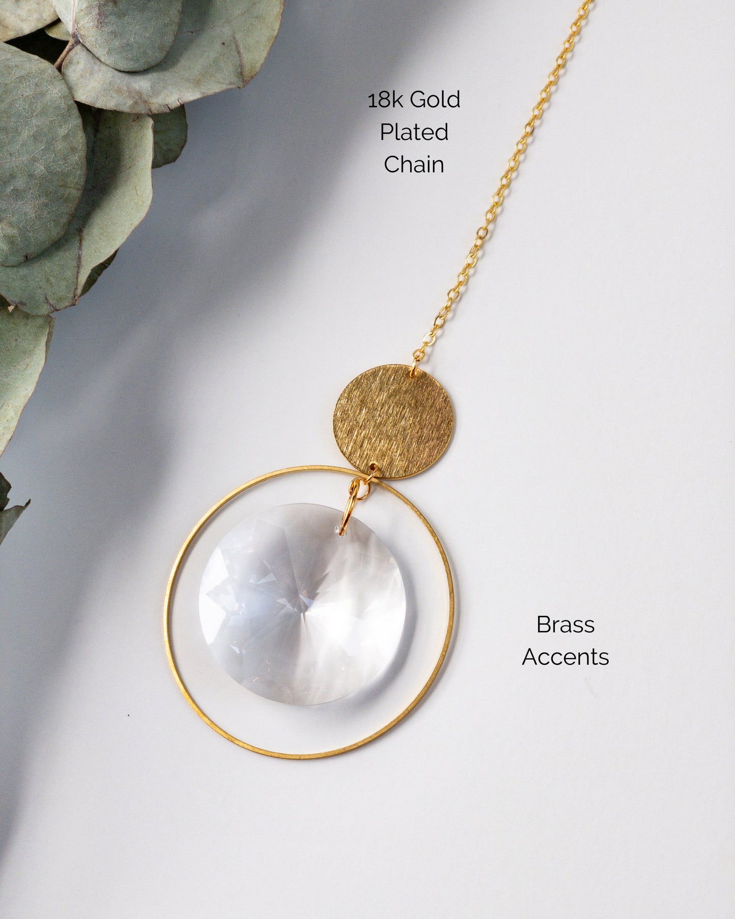 A closeup showing the materials used to make the Brass and crystal suncatcher. A brass hoop is suspended from a textured brass circle. A large disc shaped crystal is suspended in the middle of the hoop. 18k gold plated chain.