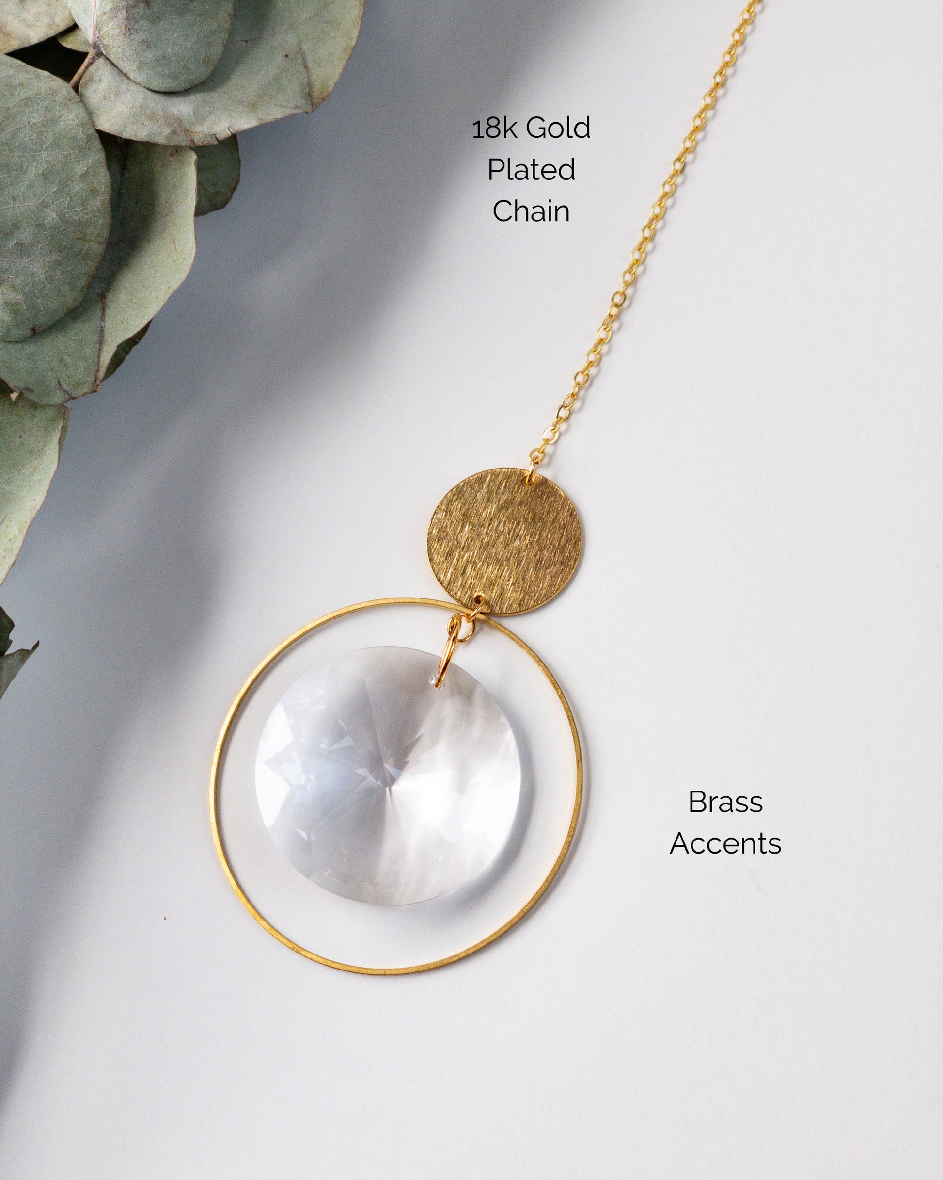 A closeup showing the materials used to make the Brass and crystal suncatcher. A brass hoop is suspended from a textured brass circle. A large disc shaped crystal is suspended in the middle of the hoop. 18k gold plated chain.