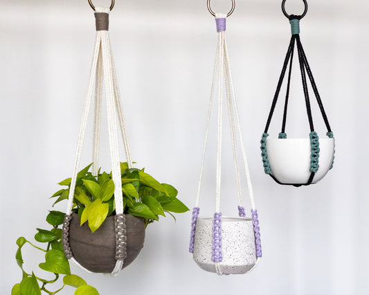 macrame plant hangers without a tassel or tail. Sleek design with 4 straight straps holding the pot. The main colors are black or white. They feature a small section along the side of the pot with an accent color in a straight knot pattern.