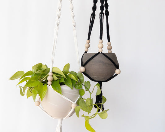 Two macrame plant hangers. One is white, one is black. Each have 3 spiral straps to hold the pots. Each strap has 3 moveable beads that sit just above the pot. Another moveable bead sits at the center of the pot. They have tassels at the bottom.