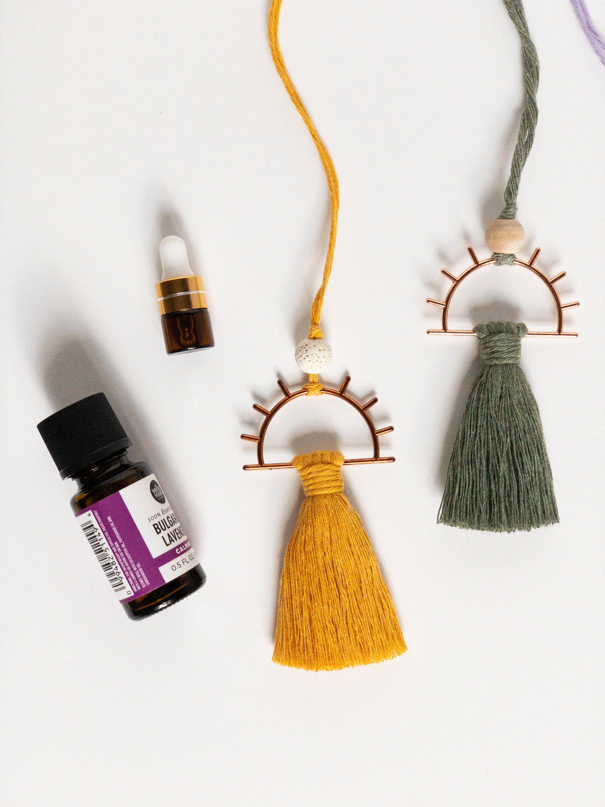 Decor piece to hang from the rearview mirror of your car. Features a metal sun with colorful brushed macrame cord tassel. A small dropper bottle of essential oils in the corner.