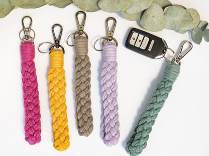 Wristlet Style Macrame keychain in a braided pattern. Shown in 5 colors, hot pink, yellow, taupe, lavender and teal. Brass, silver and smoke colored hardware. Keyfob attached to teal keychain.